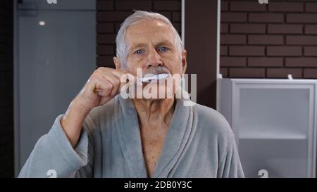 Attractive old senior man grandfather in bathrobe brushing teeth looking into mirror. Handsome elderly grandpa doing morning hygiene after shower at luxury bathroom at home. Point of view. POV shot Stock Photo