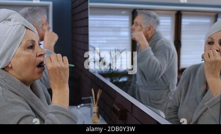 Cheerful old senior couple grandparents man and woman brushing teeth and looking into a mirror. Elderly grandmother and grandfather doing morning hygiene at luxury bathroom at home Stock Photo