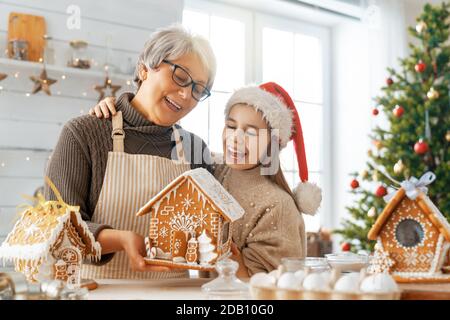 Merry Christmas and Happy Holidays. Family preparation holiday food. Grandmother and granddaughter cooking gingerbread house. Stock Photo