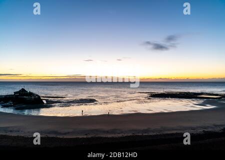 The dawn sky over the sea at low tide in a bay with beach and a single jetty harbour at one end. Coastal resort town of Broadstairs, In Kent, England. Sky is mainly blue with a thin layer of cloud and yellow on the horizon. Stock Photo