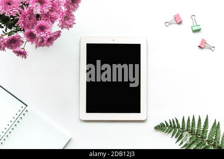 Tablet computer, notebook, paper clips, bouquet of pink chrysanthemums and fern leaves on a white background. Cozy workplace, top view, copy space. Stock Photo