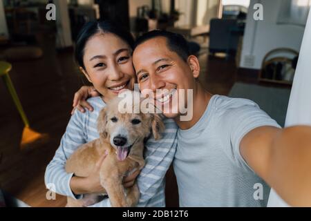 Young adult Asian couple holding a puppy taking a selfie from a phone with home interior in background. 30s mature man and woman with dog pet taking a family photo shots. - Happy group portrait Stock Photo