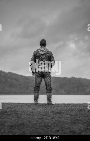 Black and white, rear view of isolated man in coat standing by a lake looking over water with moody clouds above. Stock Photo