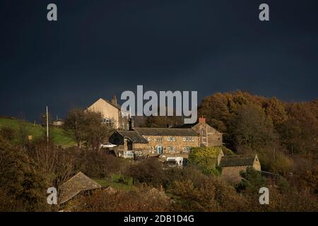 Dark black clouds gather behind some isolated stone built Yorkshire houses on a hill which are lit by the low afternoon Autumn sun near Huddersfield. Stock Photo