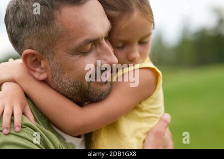 Close up of happy dad holding his lovely little daughter, smiling, standing with eyes closed while spending time together outdoors on a warm day Stock Photo