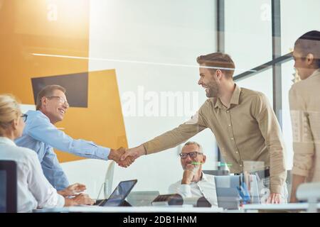 Two excited businessmen shaking hands and smiling while having a meeting with colleagues in the modern office Stock Photo