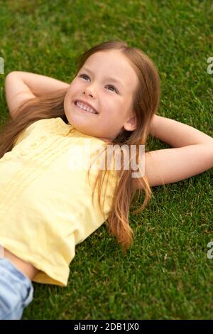 Dreaming... Portrait of joyful little girl in casual clothes lying on the green lawn, looking up and smiling while visiting park on a sunny summer day Stock Photo