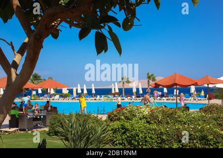 Hotel area in Egypt. Leisure beds for tourists and a cafe near the pool with blue water. Red umbrellas to protect from the sun against the blue sky in Stock Photo