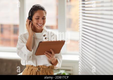Waist up portrait of smiling businesswoman wearing headset and holding digital tablet during online meeting in office, copy space Stock Photo