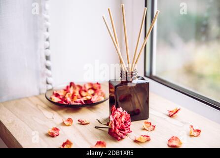 Brown glass bottle container with wood stick diffusers on home wooden window sill with beautiful dry pink rose petals for decoration. Stock Photo