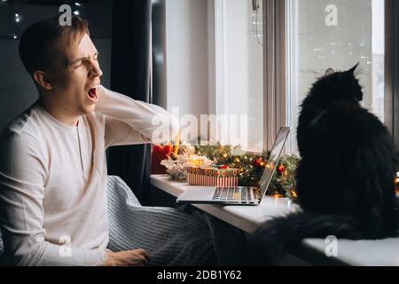 Yawning man entrepreneur in pajama tired of work online on laptop computer at home office. Christmas mood, New Year decoration, xmas vacation. Lockdow Stock Photo