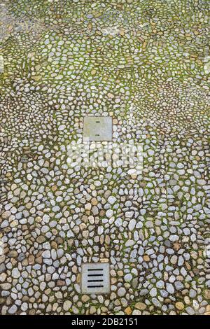 Irregular stone cobbles with moss growing between them and two metal covers in the courtyard at Rothe house, Kilkenny, County Kilkenny, Ireland Stock Photo