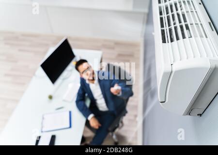 Young Businessman Using Remote Control In Front Of Air Conditioner Mounted On Wall Stock Photo