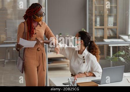 Portrait of two cheerful businesswomen wearing masks in office and bumping elbows as contactless greeting Stock Photo