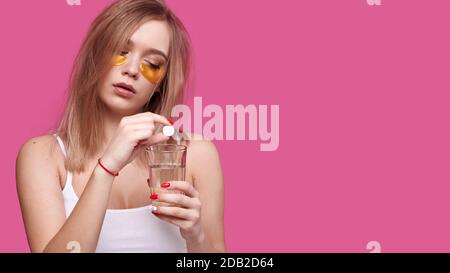 Woman with flowing hair puts pain reliever pill into glass of water after hangover on pink isolated background Stock Photo