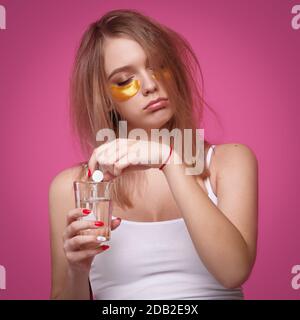 Sleepy woman with flowing hair puts pain reliever pill into glass of water after hangover on pink isolated background Stock Photo