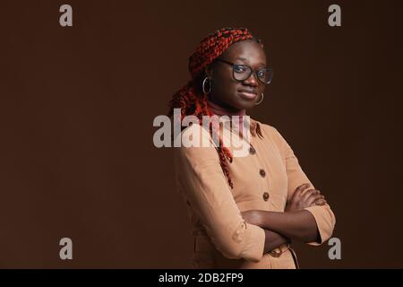 Waist up portrait of confident African-American woman looking at camera while posing standing with arms crossed against dark brown background in studio, copy space Stock Photo