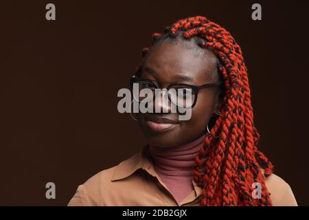 Close up portrait of confident African-American woman with braided hair smiling at camera while posing against dark brown background in studio, copy space Stock Photo