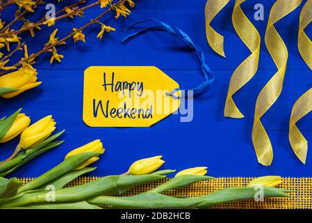 Label With English Text Happy Weekend. Yellow Spring Flowers Like Tulip And Branches. Festive Decoration With Ribbon. Blue Wooden Background Stock Photo