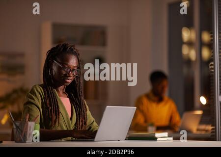 Portrait of young African-American woman wearing glasses while using laptop in office at night, copy space Stock Photo