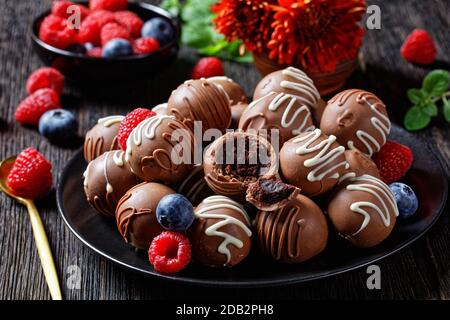 Chocolate truffles balls on a black plate on a dark wooden table with fresh berries  and flowers at the background, horizontal view from above Stock Photo