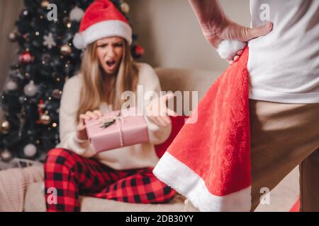 Close-up rear back view of angry man holding Santa hat and unhappy young woman with Christmas present box near Christmas tree. Couple problems concept Stock Photo
