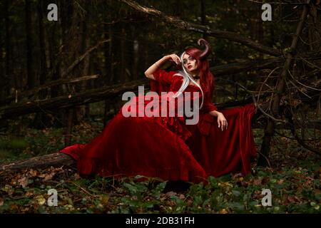 Maleficent Woman in Red Clothing and Horns in dark Forest. Posing in magik forest Stock Photo