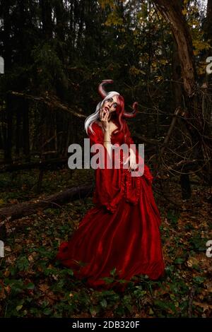 Maleficent Woman in Red Clothing and Horns in dark Forest. Posing in magik forest Stock Photo