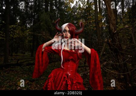 Maleficent Woman in Red Clothing and Horns in dark Forest. Posing in magik forest Stock Photo