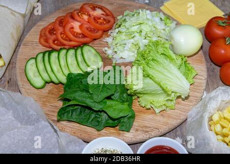 Preparing for the preparation of hamburgers. On a wooden board are lettuce leaves, chopped: tomatoes, cucumbers, onions, cutlets and sauces. Stock Photo