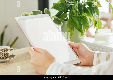 Tablet white screen mockup held by a woman's hand who is in the living room. Blank screen to insert design. Stock Photo
