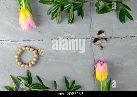 Jewelry, bracelet with keychain and gold earrings together with tulips and green laid on a concrete background. Inside the composition, free space. Stock Photo