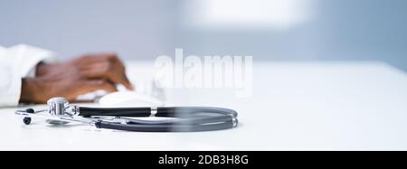 View Of Stethoscope With Doctor Using Computer Keyboard In Clinic Stock Photo