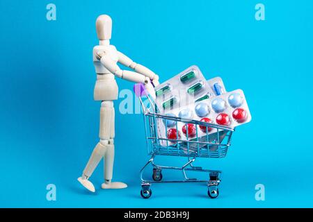 Wooden doll and metal shopping cart with pills on blue background. Various capsules, tablets and medicine on blue pastel trendy background. Pills conc Stock Photo