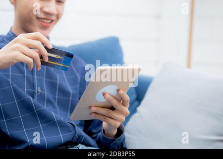 Young asian man smiling holding credit card shopping online with tablet computer buying and payment, male using debit card purchase or transaction of Stock Photo