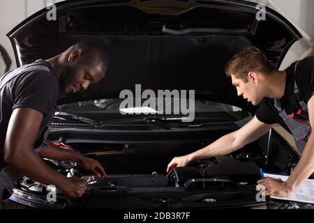 two diverse auto mechanics cooperating while examining car engine performance in auto repair shop, work together, hold diagnostics Stock Photo