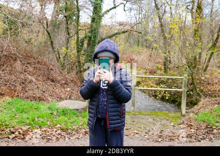 Child boy 10 or 11 taking photo holding a mobile phone (cell phone) while on a walk in autumn wearing a hoodie and winter jacket Wales UK KATHY DEWITT Stock Photo