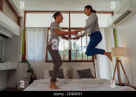 Portrait of happy young adult Asian couple jumping on bed together in bedroom interior scene. 30s mature husband and wife smiling and holding hands. Marriage and happy relationship life concept Stock Photo