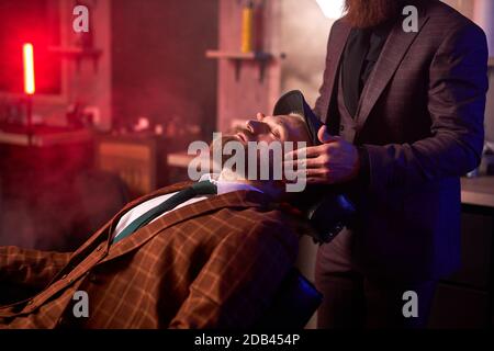 young caucasian handsome man on chair of barber having procedure of shaving, get beauty procedures in barber shop salon Stock Photo