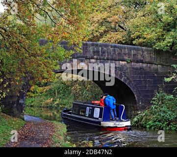 A couple share a kiss on a canal Narrowboat as it passes under a stone bridge in the autumn colours  on the Leeds and Liverpool canal near Adlington Stock Photo