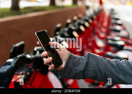 detail of a hand with a smart phone next to an urban parking of rental bikes, concept of technology and sustainable mobility Stock Photo