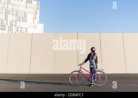 young girl with a pink retro bike at city adjusting her protective mask, concept of active lifestyle, protection against covid and sustainable urban m