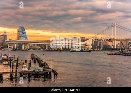 Sunset on the circular highway leading to the Rainbow Bridge with Cargo and cruise ships moored or sailing in Odaiba Bay of Tokyo. Stock Photo