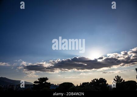 Sun hiding behind a cloud, blue cloudy sky over trees and buildings near hill in town Stock Photo
