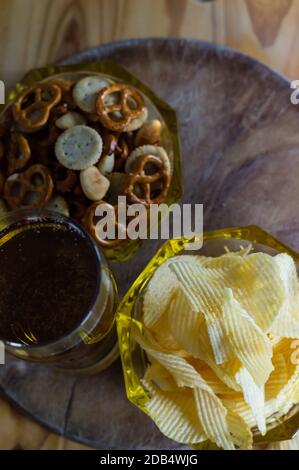 Cold beer and wavy potato chips in a bowl and salty snacks pretzel on a wood table from above Stock Photo