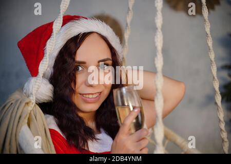 Young woman in Santa Claus suit with glass of champagne sitting on hammock. Attractive female in Christmas hat swinging on hammock chair. Concept of C Stock Photo