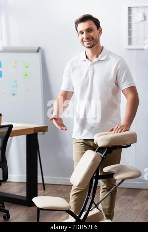 Front view of cheerful masseur standing and pointing with hand at massage chair in office Stock Photo