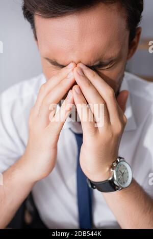 Close up view of tired businessman with headache, holding hands near closed eyes on blurred background Stock Photo