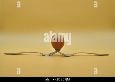 egg balanced on a composition of 2 intertwined forks isolated over neutral orange background Stock Photo