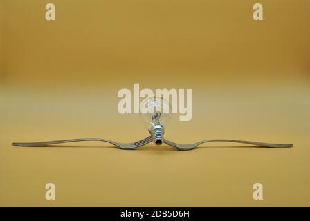 light bulb balancing on composition of 2 intertwined forks isolated on orange neutral background, conceptual photo to indicate division of electricity Stock Photo